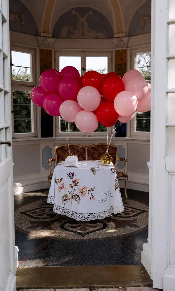 Ein liebevoll gedeckter Tisch mit Stickerei und rosa-roten Ballons steht in einem lichtdurchfluteten Raum mit Rundbogenfenstern.