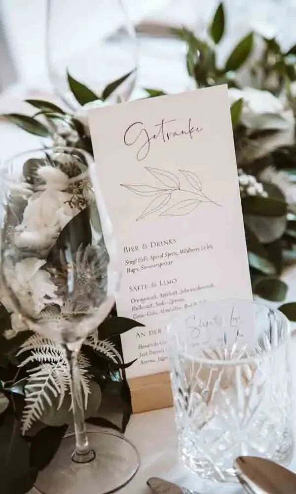 Elegante Getränkekarte mit Blättermotiv, eingerahmt von Gläsern, weißer Tischdeko und frischen Grünakzenten.