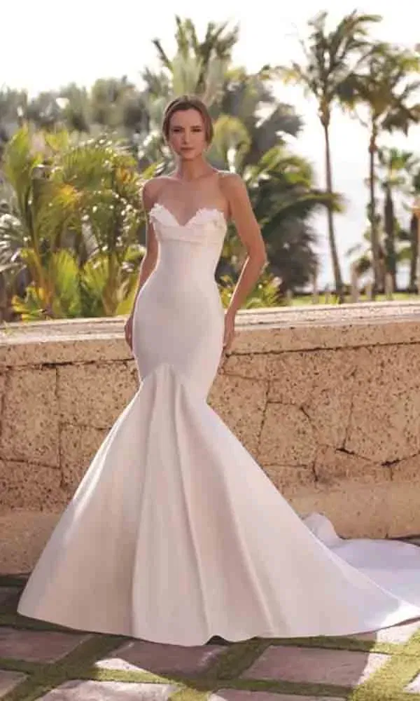 Elegantes Mermaid-Brautkleid mit figurbetontem Schnitt und dezentem Spitzenbesatz, inszeniert im sonnigen Außenbereich.