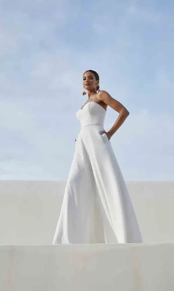 Moderner Hochzeitslook: Frau im weißen trägerlosen Jumpsuit mit weitem Schnitt auf heller, minimalistischer Kulisse.
