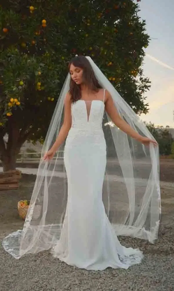 Schlichte, elegante Braut in figurbetontem Kleid mit langer Schleppe posiert bei Sonnenuntergang unter Obstbäumen.