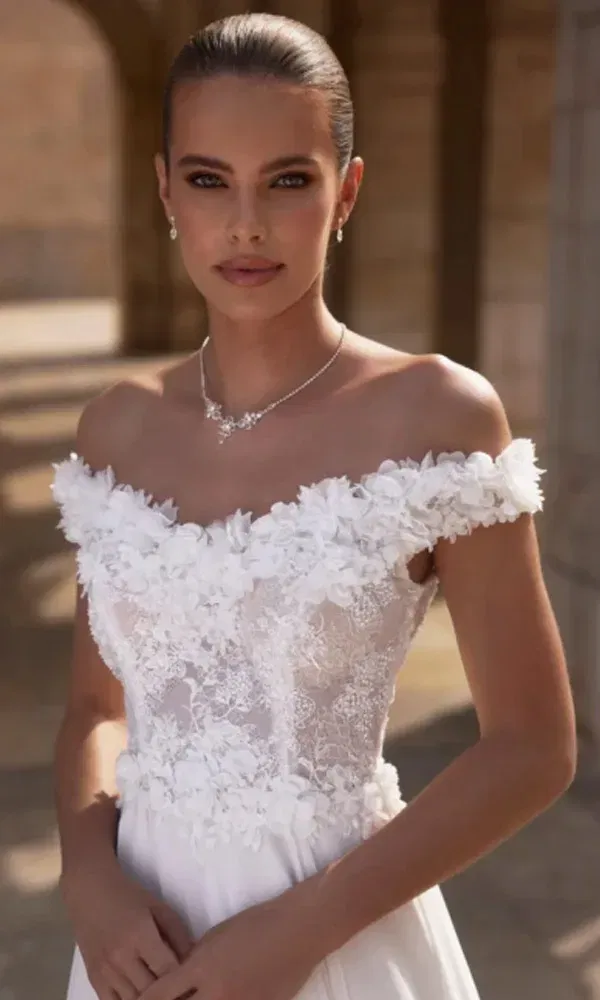 Nahaufnahme einer eleganten Braut im trägerlosen Spitzenkleid mit 3D-Blüten und zartem Schmuck im Sonnenschein.