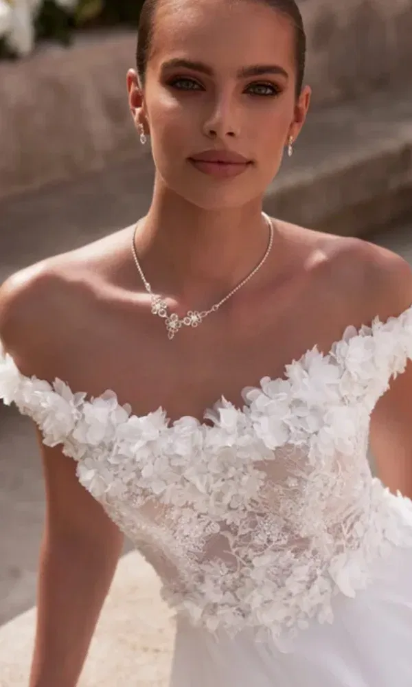 Zarte Spitze mit 3D-Blüten: Braut sitzt in einem romantischen Hochzeitskleid auf einer sonnigen Sandsteintreppe.