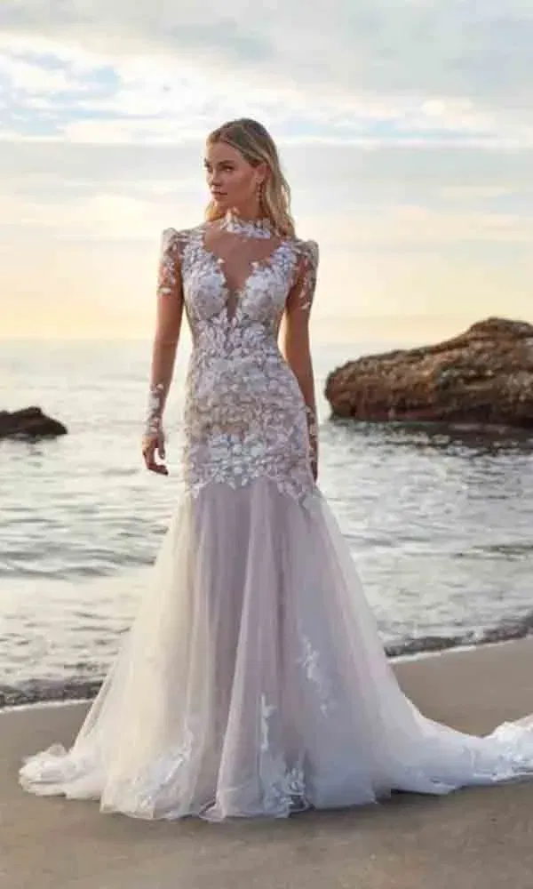 Transparentes Brautkleid mit floraler Spitze, getragen von einer Braut am Meer – dramatische Kulisse und elegante Pose.