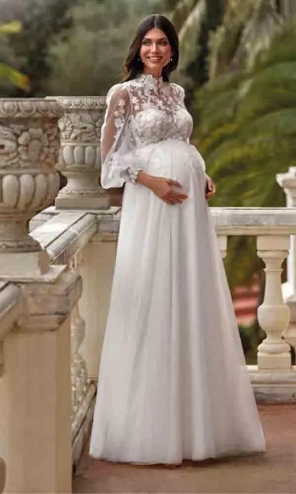 Schwangerschaftsbrautkleid mit Stehkragen und Spitzeneinsatz, getragen von einer lächelnden Braut vor Palmenkulisse.