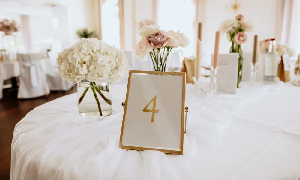 Festlich gedeckter Hochzeitstisch mit goldgerahmter Tischnummer, Blumen in Weiß und Rosé sowie stilvollen Kerzenhaltern.