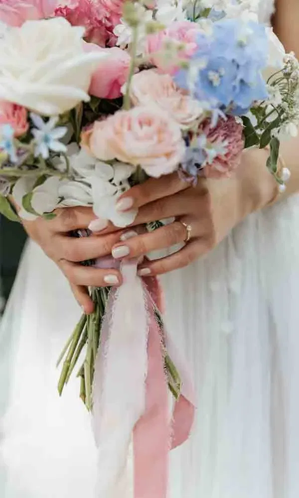 Detailfoto von Händen mit Verlobungsring, die einen bunten Hochzeitsstrauß mit rosafarbenen Bändern festhalten.
