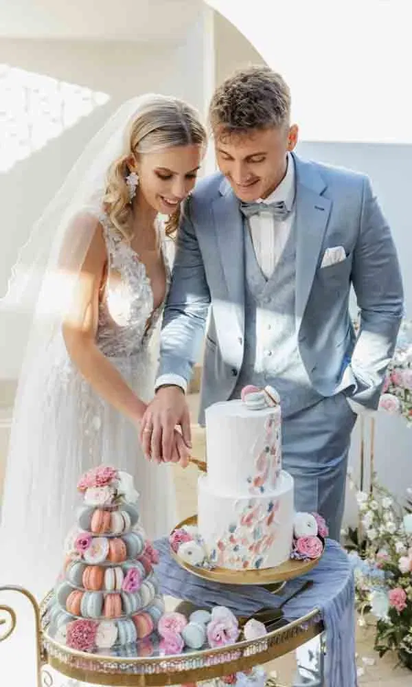 Bräutigam im blauen Anzug und Braut im Spitzenkleid beim Tortenanschnitt – elegante Torte und farbenfrohes Sweet Table-Arrangement.