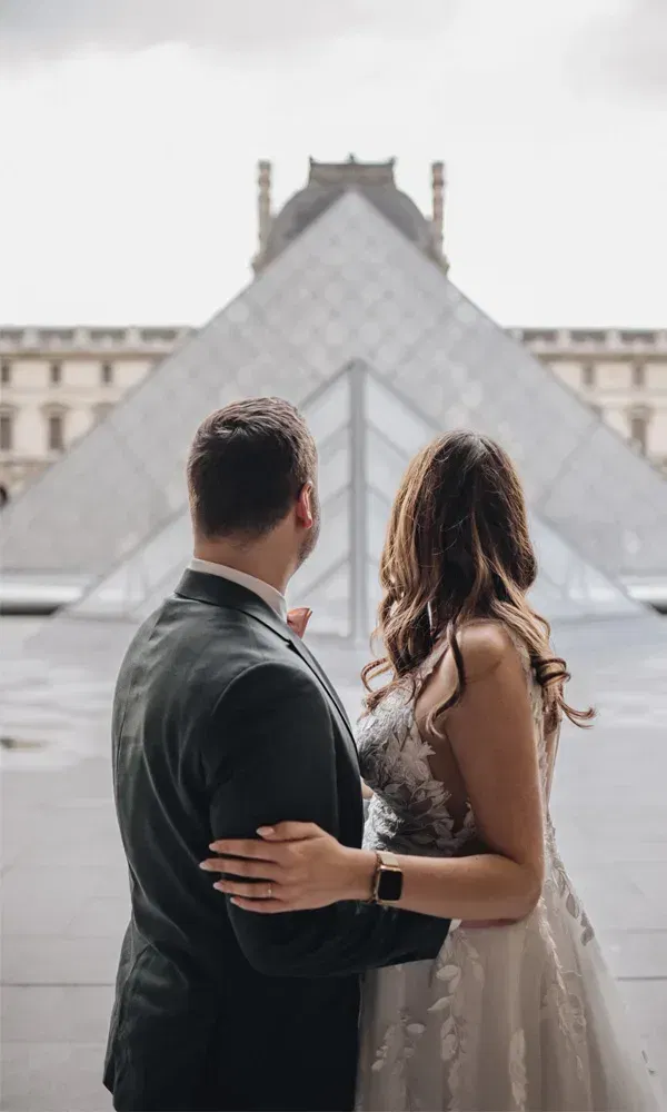 Rückansicht eines Hochzeitspaares vor dem ikonischen Louvre-Gebäude mit moderner Glaspyramide.