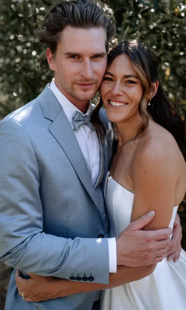 Braut in trägerlosem Kleid und Bräutigam im hellblauen Anzug umarmen sich liebevoll und blicken lächelnd in die Kamera.