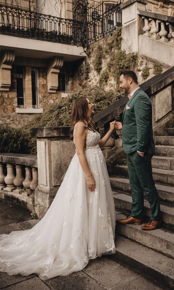 Braut in Spitzenkleid und Bräutigam im grünen Anzug beim liebevollen Moment auf Steinstufen.