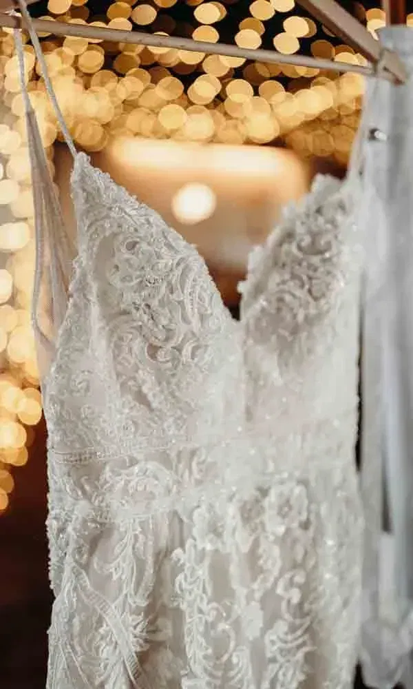 Weißes Hochzeitskleid mit feiner Spitze auf einem Kleiderbügel, im Hintergrund goldenes Bokeh von Lichterketten.