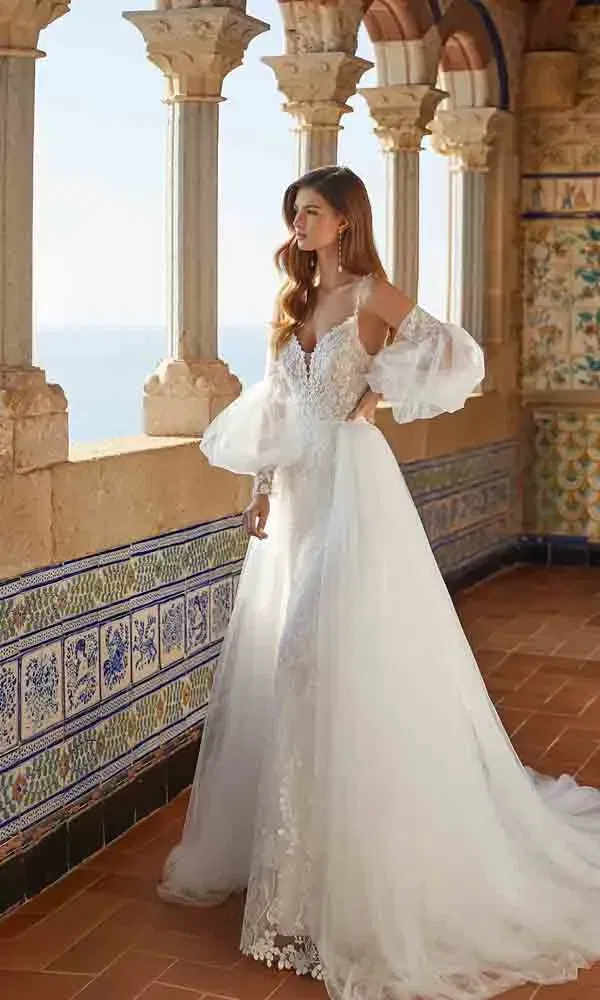 Romantisches A-Linien-Brautkleid mit transparenten Statement-Ärmeln und tiefem Ausschnitt vor einer kunstvollen Säulenkulisse.