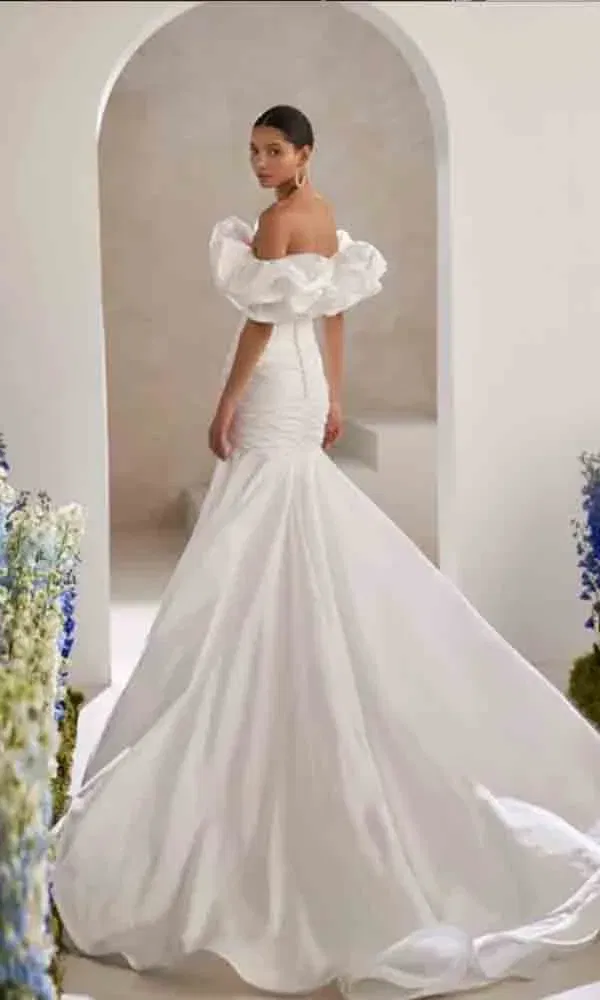 Elegante Braut in moderner Robe mit voluminösen Ärmeldetails und geschwungener Silhouette, aufgenommen in einem minimalistischen, blumengeschmückten Innenraum.