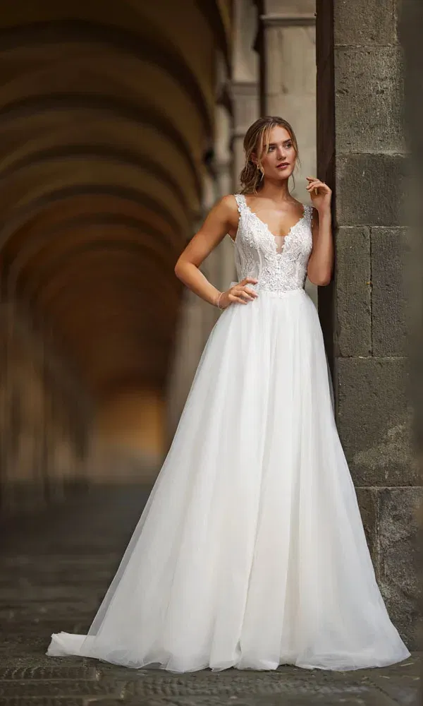 Romantisches Brautkleid mit floral besticktem Mieder und fließendem Tüllrock – die Braut steht nachdenklich an einem Steinpfeiler.