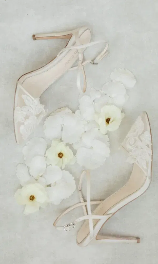 Hochzeitsschuhe mit feiner Spitze und femininen Blüten, flach auf hellem Untergrund arrangiert.
