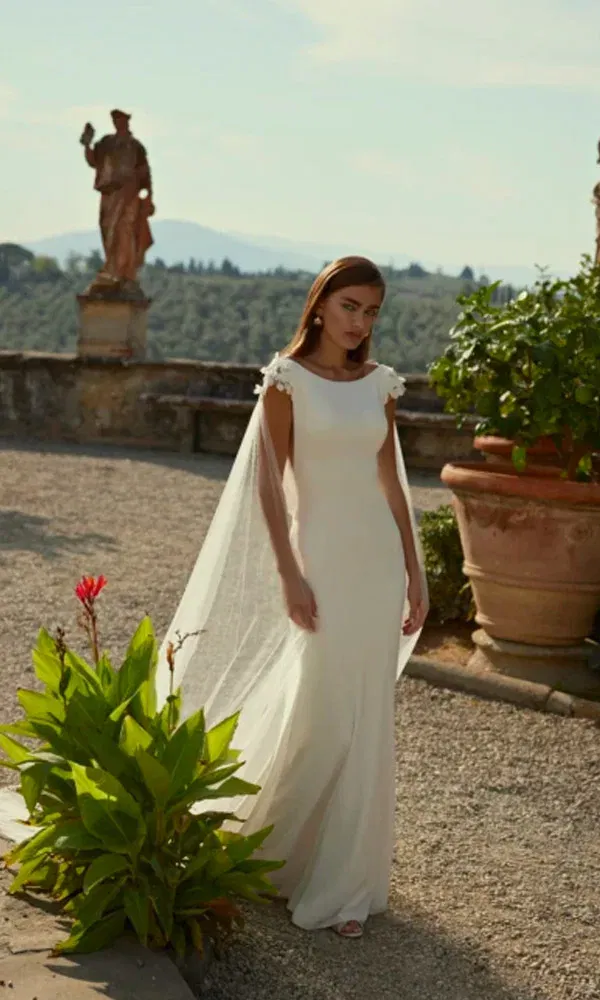 Ganzkörperaufnahme einer Braut mit cleanem Cape-Kleid und floralen Schulterdetails im historischen Garten.