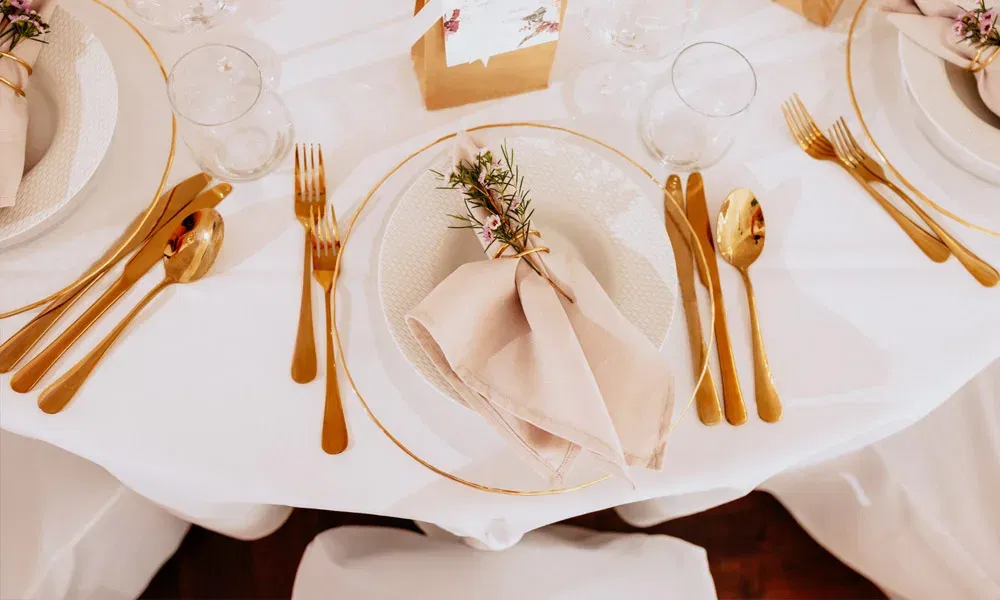 Stilvolles Hochzeitsgedeck mit goldenen Besteckdetails, gefalteter Stoffserviette und liebevoller Kräuterverzierung.