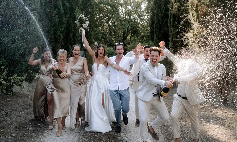 Jubel, Sekt und Sommerfreude: Das Brautpaar zieht mit Freunden lachend durch die Natur, während der Sekt in die Luft spritzt.