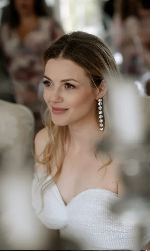 Die Braut sitzt strahlend in einem hellen Raum, trägt ein weißes Off-Shoulder-Kleid und auffällige Ohrringe.