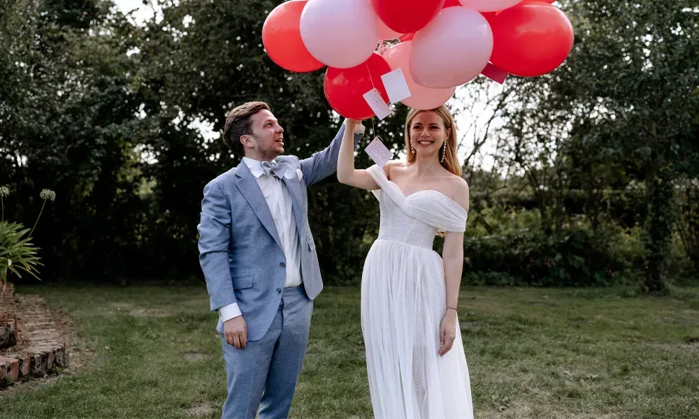 Fröhliche Szene im Grünen: Die Braut hält Ballons mit Grußkarten, der Bräutigam im blauen Anzug schaut sie bewundernd an.