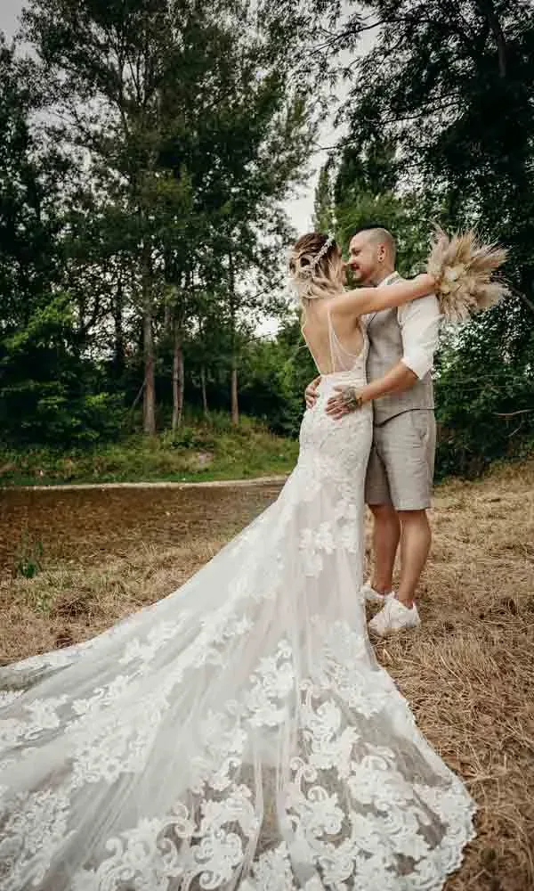 Intimer Moment in der Natur: Die Braut mit Trockenblumenstrauß und langer Spitze, der Bräutigam in Shorts – beide umarmen sich liebevoll.