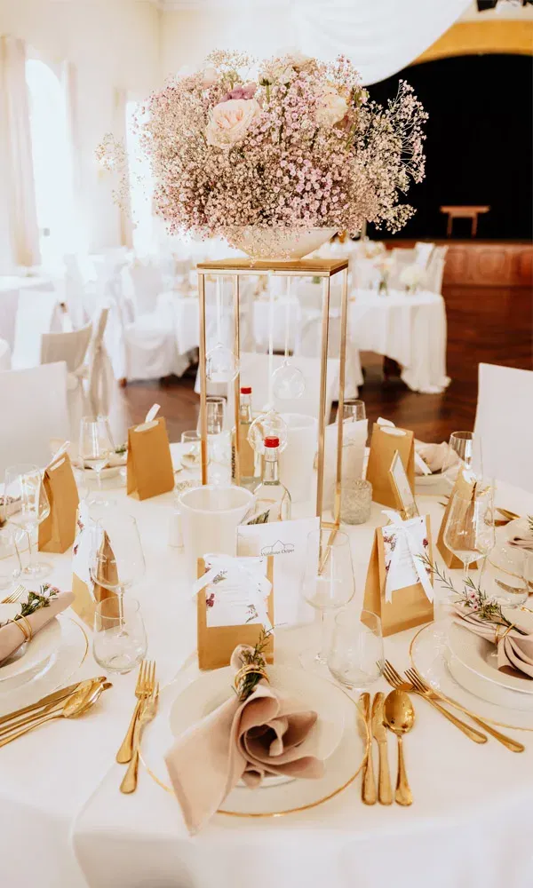 Eleganter Hochzeitstisch mit hoher Blumenschale aus zartem Rosa und Weiß, goldenen Bestecken und liebevollen Details.