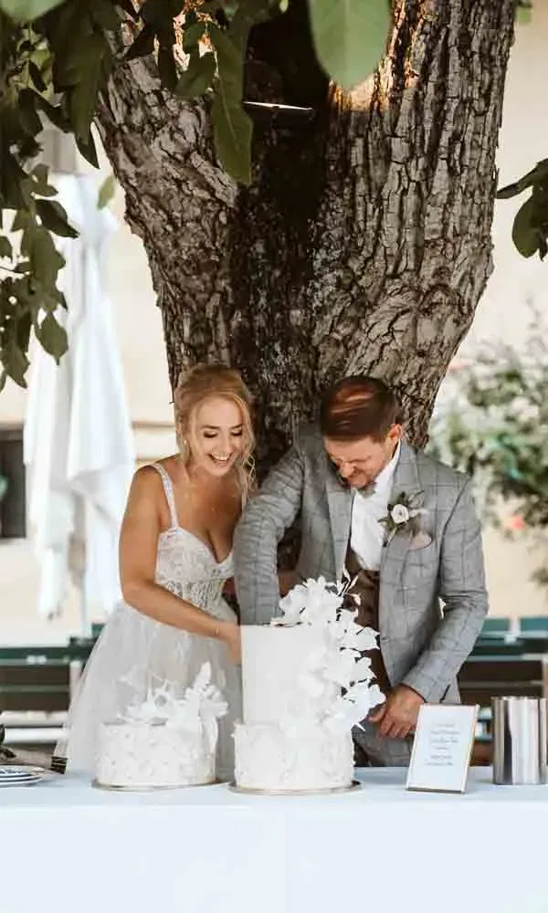 Sophie und Reinhard beim traditionellen Anschnitt der zweistöckigen Torte mit weißen Blüten im Freien.