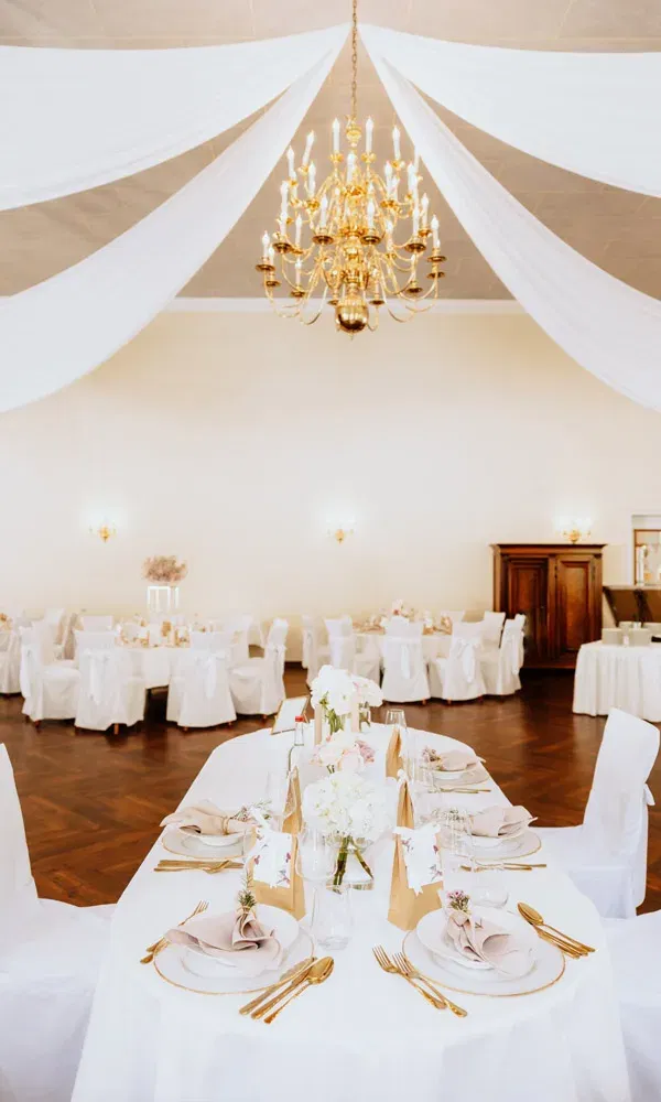 Stilvoll dekorierter Hochzeitssaal mit goldenen Bestecken, Blumenschmuck und einem großen Kronleuchter unter weißen Stoffbahnen.