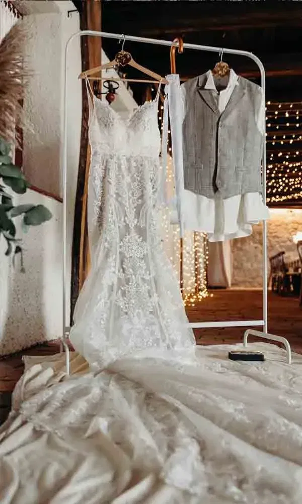 Romantisches Getting-Ready-Foto: Spitzenkleid und Anzugweste auf einem weißen Kleiderständer vor stimmungsvoller Hochzeitsdeko.