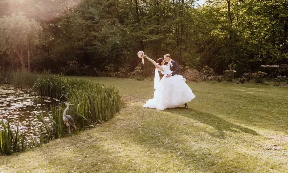 Romantische Szene bei Sonnenuntergang: Das Brautpaar posiert tanzend auf einer Wiese am Teich, ein Reiher beobachtet das Geschehen.