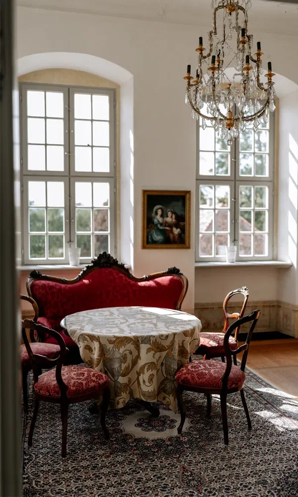 Stilvoll eingerichteter Raum mit antikem Mobiliar, großen Fenstern und einem Gemälde an der Wand.