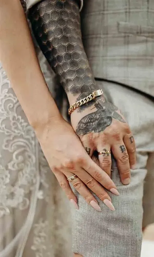 Detailfoto: Zwei Hände mit Eheringen ruhen übereinander – sichtbar sind Tattoos, Schmuck und feine Stoffdetails vom Hochzeitskleid.