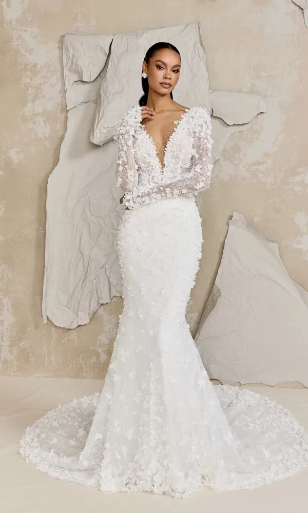 Model trägt ein aufwendig verziertes Brautkleid mit V-Ausschnitt, langen Ärmeln und floralen Applikationen, posierend vor abstrakter Wanddeko in einem Atelier-Setting.