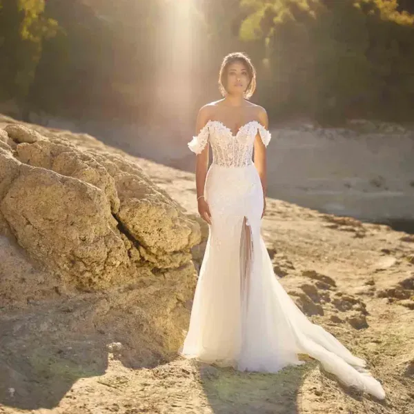 Brautmodell in figurbetontem Brautkleid mit Carmen-Ausschnitt und langer Schleppe steht in dramatischer Naturkulisse bei Sonnenuntergang.