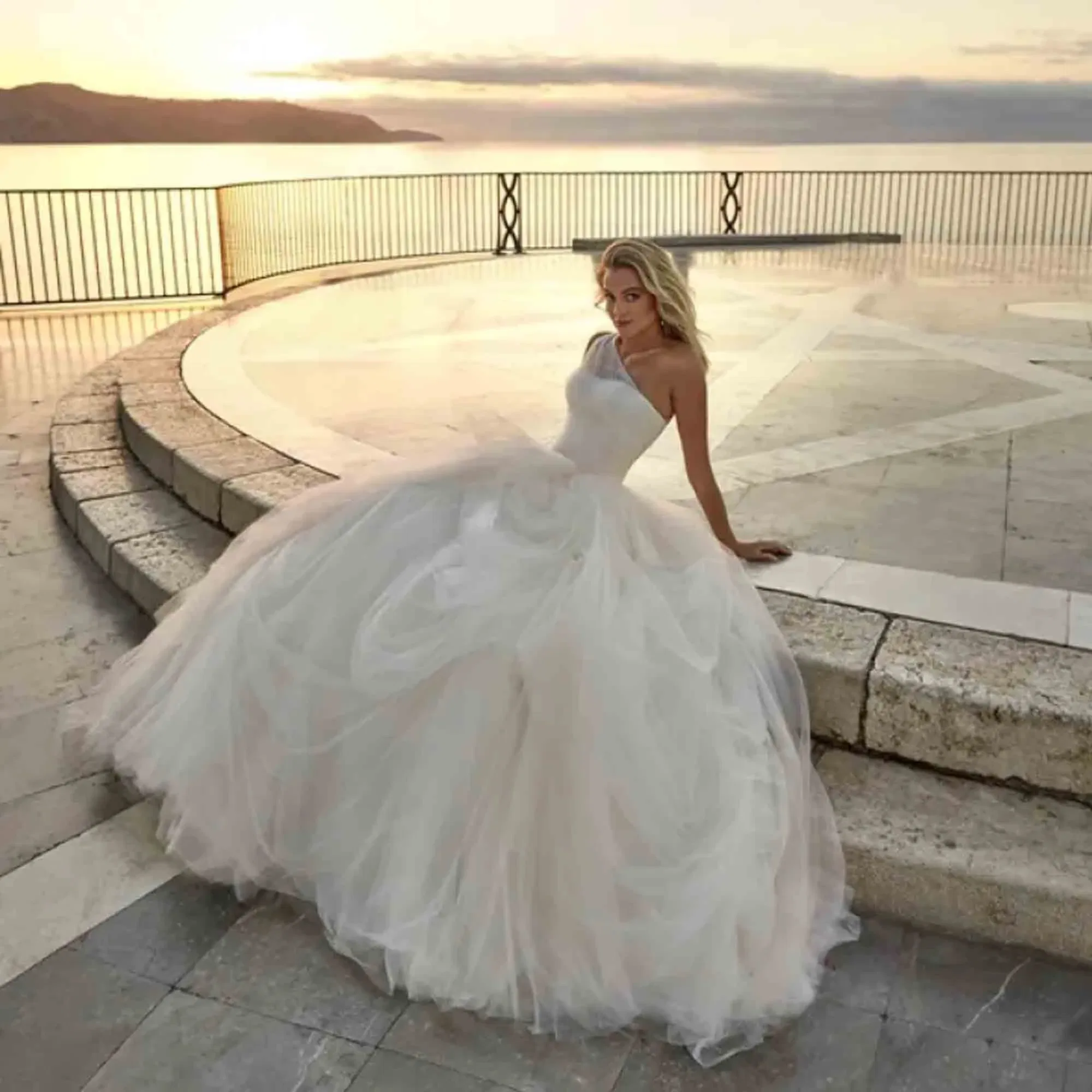 Stilvolle Braut in einer traumhaften Prinzessinnenrobe mit voluminösem Rock posiert an einer marmorglänzenden Uferpromenade bei Abendlicht.