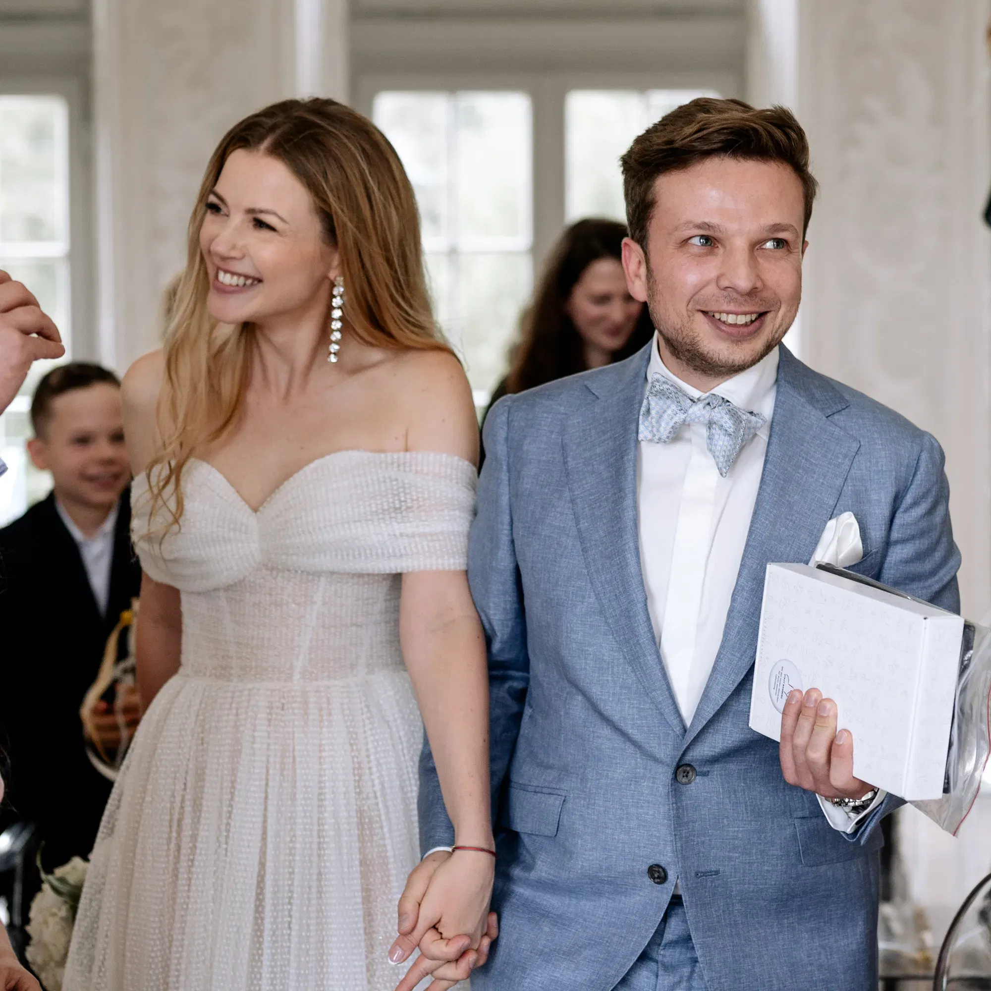 Ein frisch verheiratetes Paar verlässt gemeinsam den Trausaal, die Braut im schulterfreien Kleid, der Bräutigam mit Geschenk in der Hand.