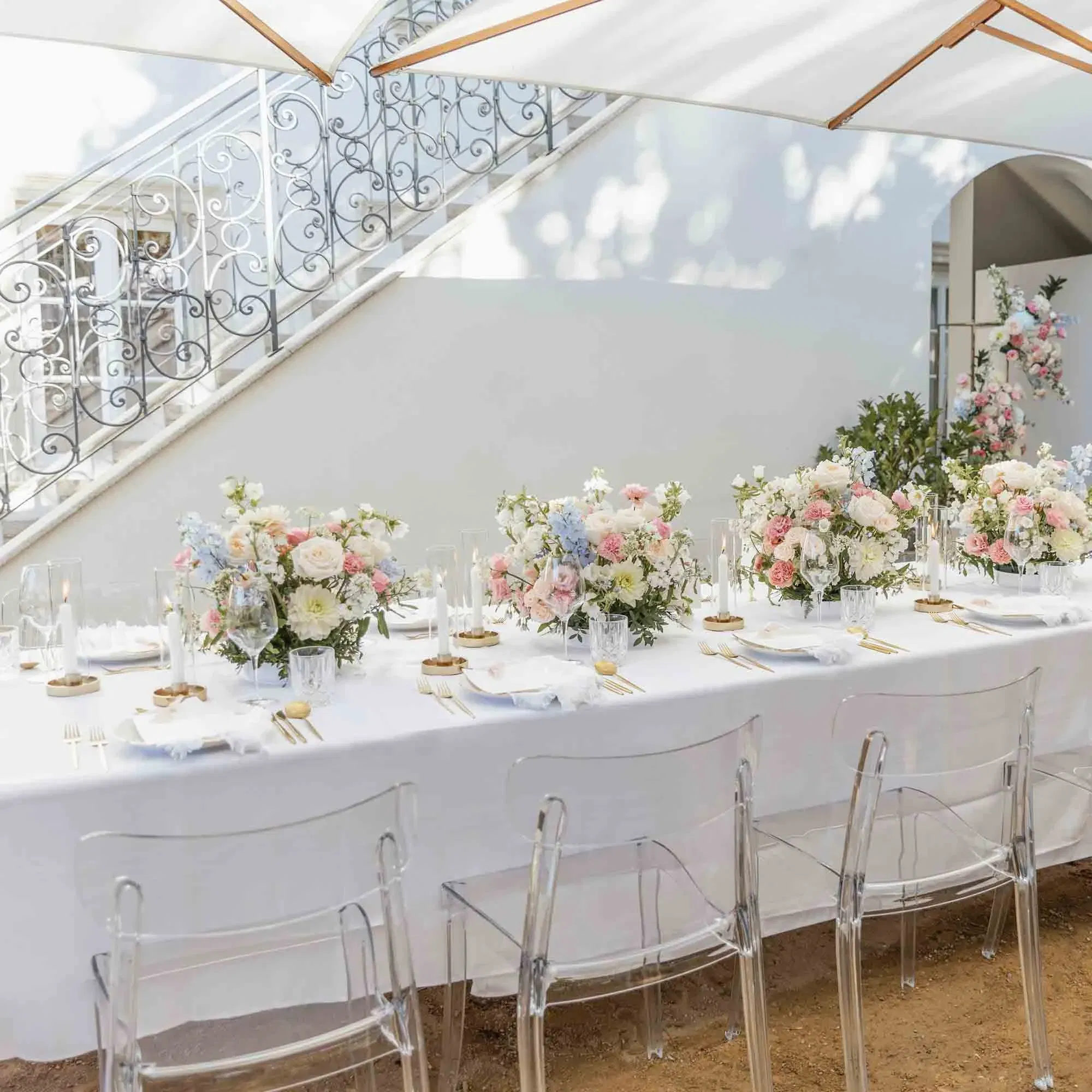 Outdoor-Hochzeitsessen: stilvoll gedeckter Tisch mit zarten Blumenarrangements in Rosé und Hellblau, umgeben von transparenten Stühlen.