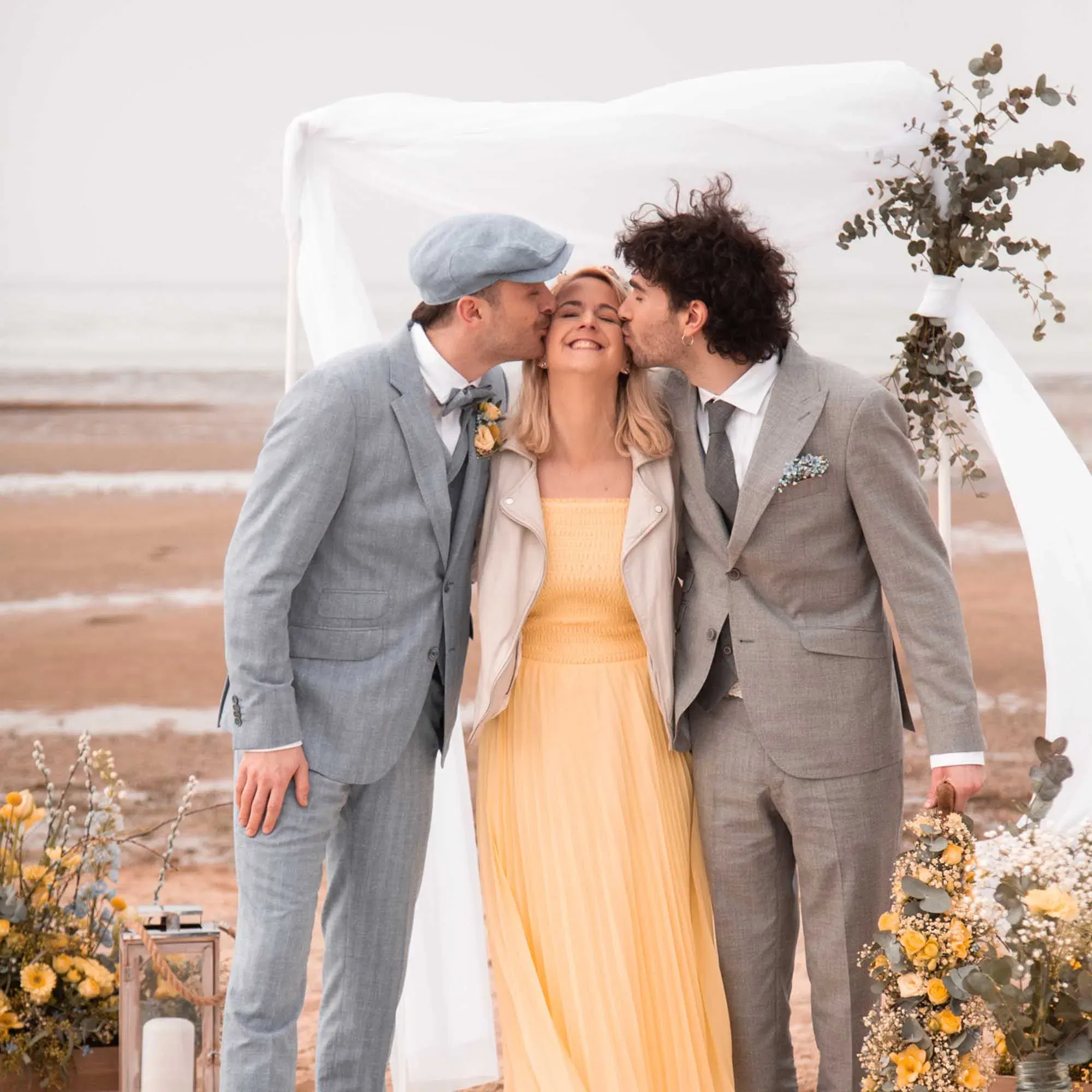 Herzlicher Moment am Meer: Ein Hochzeitspaar mit Begleitung feiert eine inklusive Trauzeremonie am Strand, eingerahmt von weißen Stoffen und gelber Blütenpracht.
