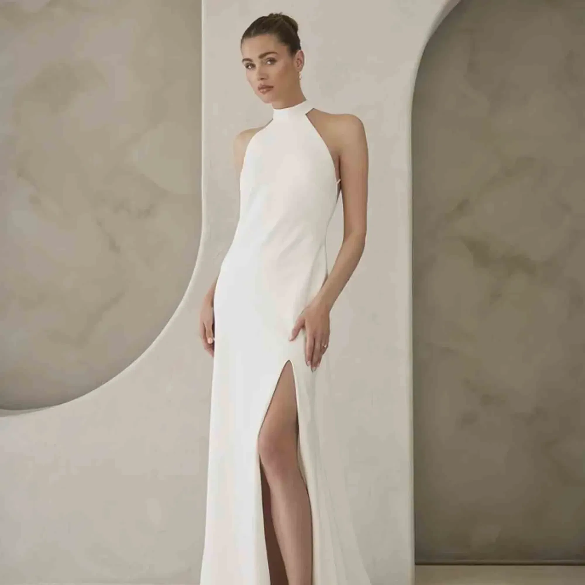 Elegantes Etui-Brautkleid mit Halterneck und hohem Schlitz – fotografiert vor hellem, architektonischem Hintergrund.