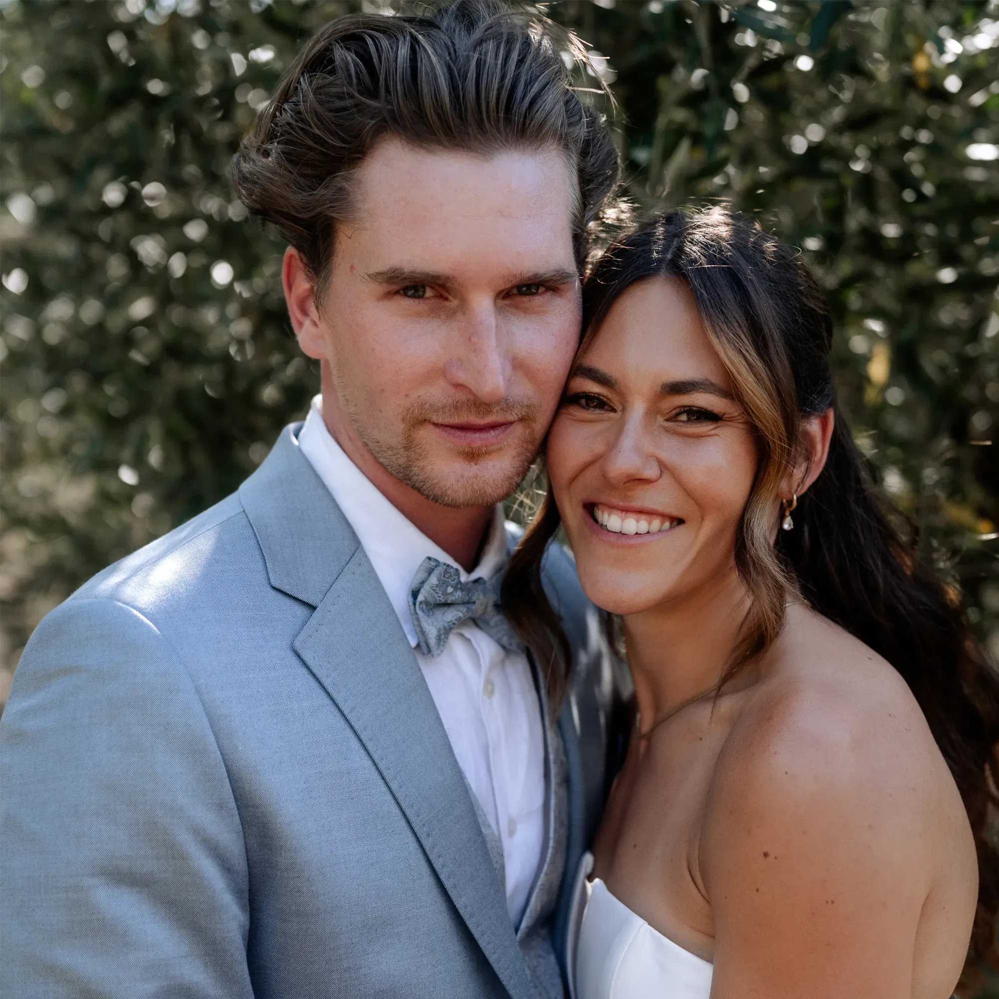 Frisch vermähltes Paar strahlt bei einer Hochzeit im Grünen – er im hellblauen Anzug, sie im schulterfreien Brautkleid.