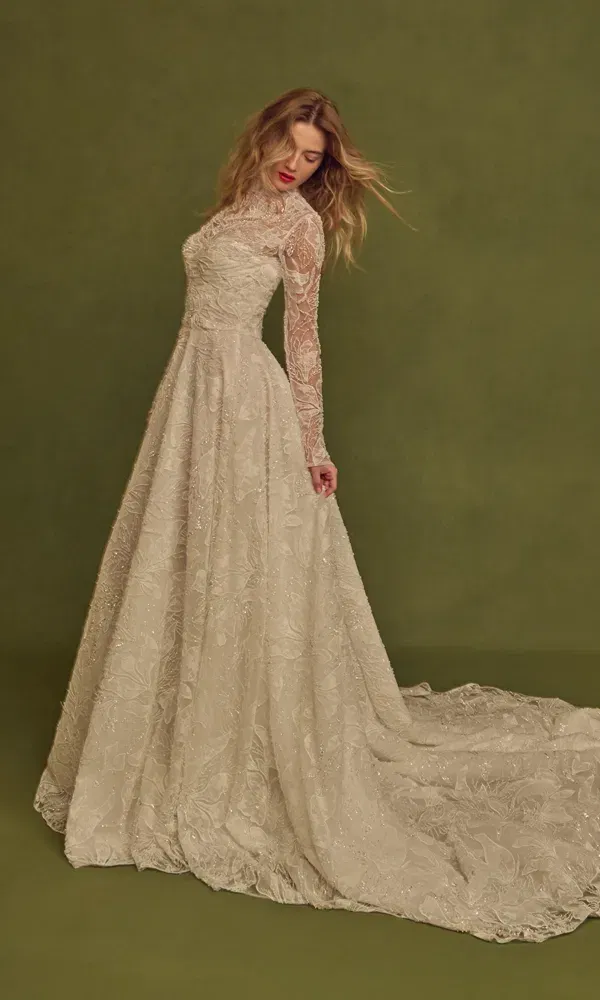 Model trägt ein elegantes, vintage-inspiriertes Brautkleid mit Stehkragen, transparenten Ärmeln und zarter Stickerei, fotografiert vor moosgrünem Hintergrund.