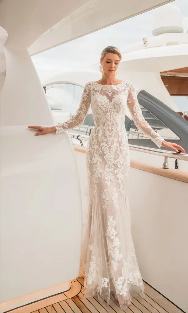 Model trägt ein zart besticktes Hochzeitskleid mit langen Ärmeln und Bootsausschnitt, fotografiert auf einem Schiff mit Blick auf futuristische Linien und Meerblick.