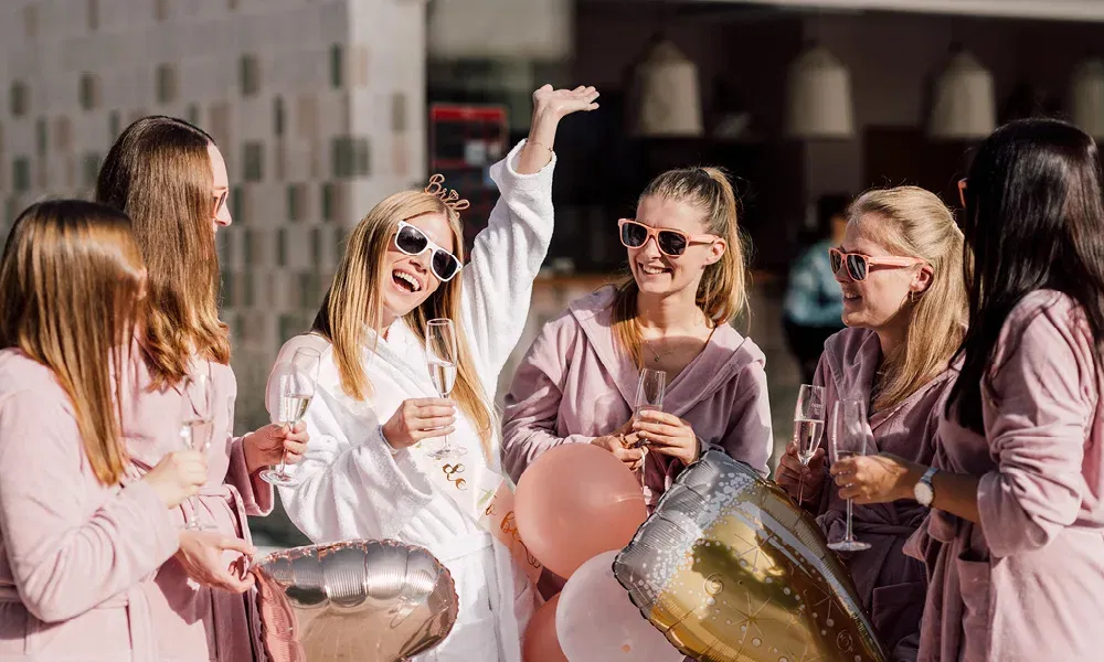 Die Braut in weißem Bademantel feiert mit ihren Freundinnen bei Sonnenschein, alle tragen Sonnenbrillen und stoßen mit Sekt an.