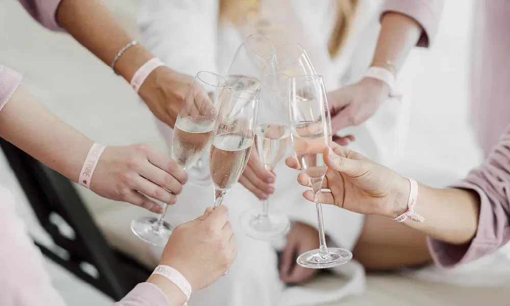 Sechs Freundinnen stoßen mit Sektgläsern an, ihre rosa Armbänder zeigen den Schriftzug „Bride Tribe“.