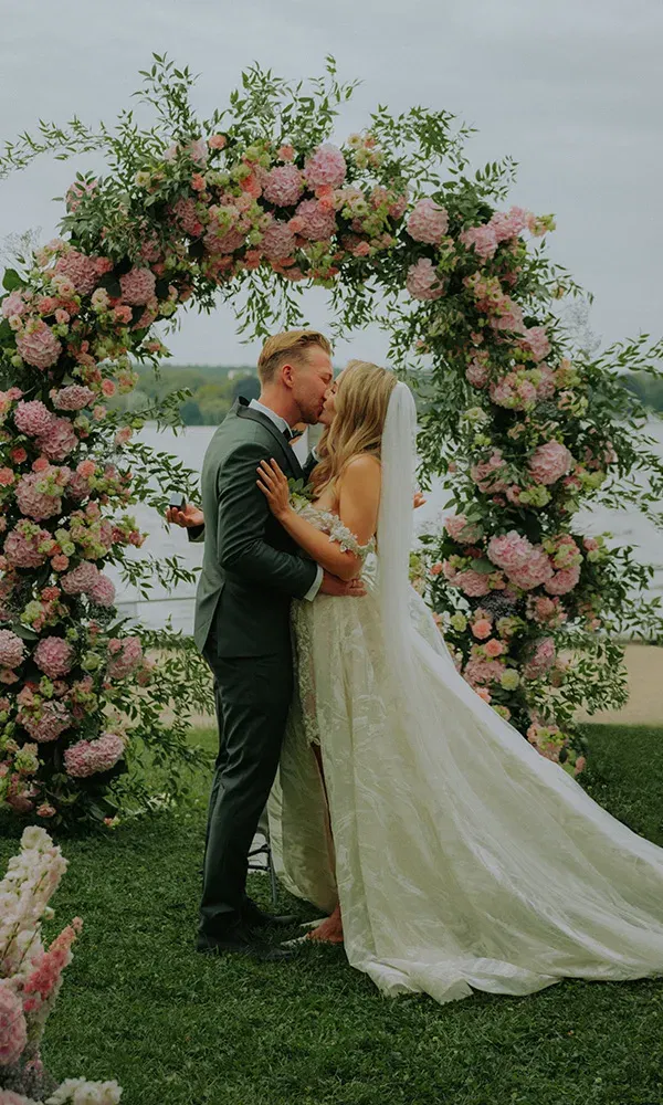 Romantischer Hochzeitskuss am See unter einem Blütenbogen in Rosa und Grün.