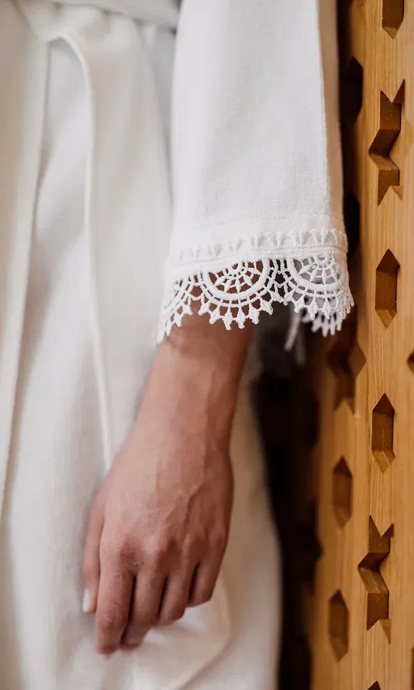 Nahaufnahme eines weißen Bademantels mit zarter Spitzenborte am Ärmel, daneben eine Hand an einer Holzwand mit Sternenmuster.