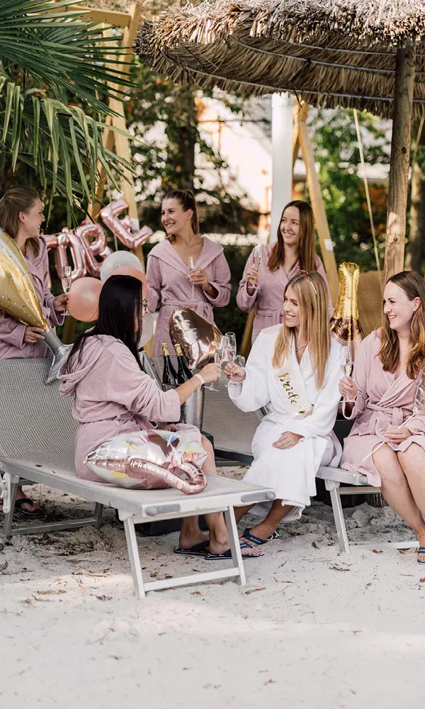 Die Braut und ihre Freundinnen sitzen im Sand unter Palmen und stoßen auf ihren JGA an – mit Luftballons und Sekt in der Hand.