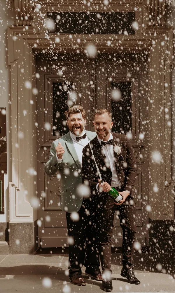 Frisch verheiratetes Paar stößt vor einer Location mit Champagner an, umgeben von prickelnden Tropfen.