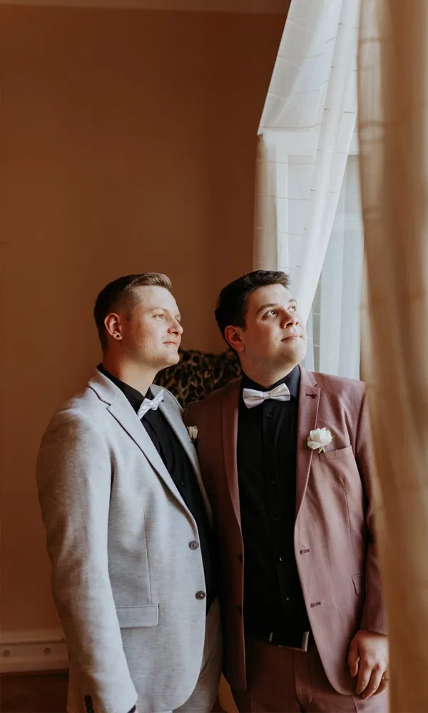 Hochzeitspaar in eleganten Anzügen blickt seitlich aus dem Fenster, sanftes Licht fällt durch den Vorhang.